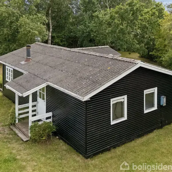 En sortmalet træhytte står stille i en grøn have omkranset af træer. Hytten har en veranda med et hvidt rækværk og en skorsten på taget.