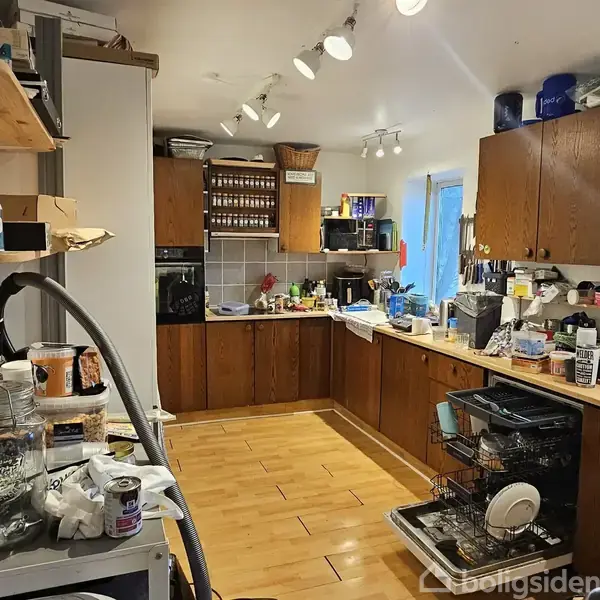 Køkken med træskabe, opvaskemaskine fuld af service, borde oversået med diverse køkkenartikler og madlavningselementer. Hylder til venstre opbevarer krukker og rengøringsmidler. Lyse trægulve og hvidt loft.