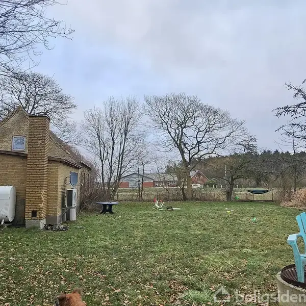 En lille, gul murstensbygning står i en have med græs og spredte træer. En blå stol og et bord er i nærheden, og landskabet er åbent med flere træer i baggrunden.