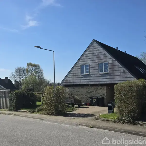 En trekantet tagdækket hus står stille på en rolig vej, omgivet af hække og en forhave. En enkelt gadelampe kaster skygger over asfalten.