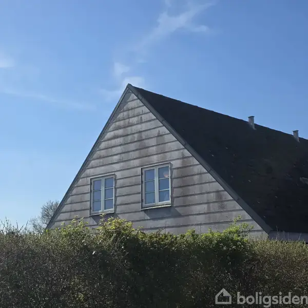 Et trekantet hus med et mørkt tag og to vinduer er omgivet af grønne buske under en klar blå himmel.