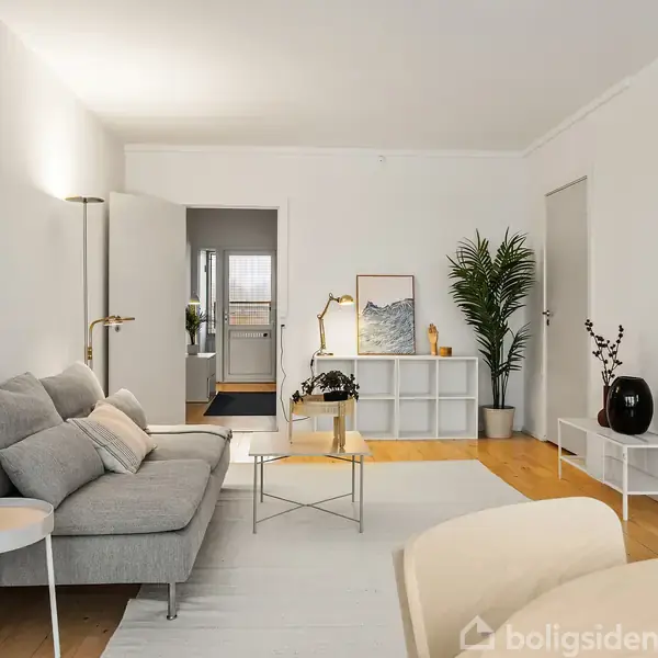 Grå sofa står i en lys stue ved siden af et lille, rundt bord. En stor plante og kunst på en hylde dekorere rummet. Trægulv og lyse vægge omkranser det hele.