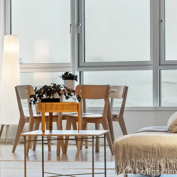 Et lille spisebord med fire stole står ved siden af store vinduer i en lys stue; en plante og en lampet lyser op ved siden af sofaen.