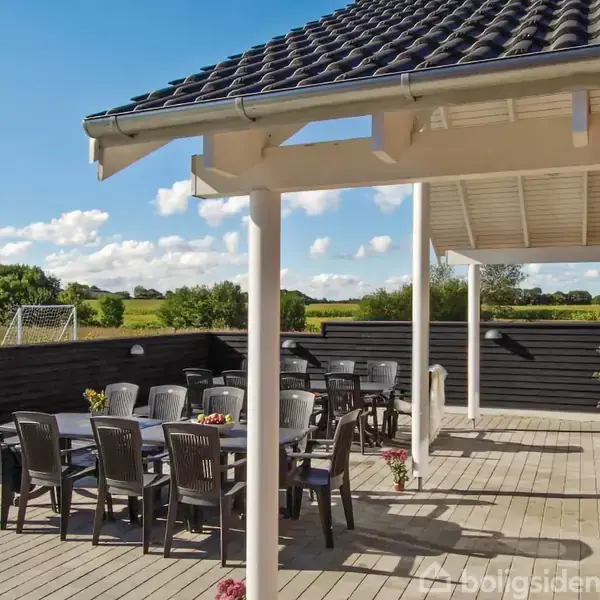Terrasse med sortmalede træmøbler arrangeret omkring flere borde, omkranset af sorte træhegn. I baggrunden ses en græsplæne med et fodboldmål under en klar blå himmel.