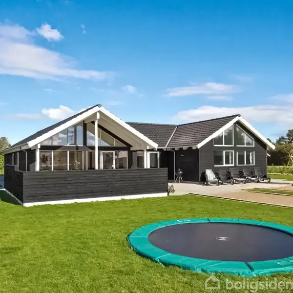 Et sort træhus med store vinduer står på en grøn græsplæne. I baggrunden ses en legeplads og en trampolin. Himlen er blå med enkelte skyer.