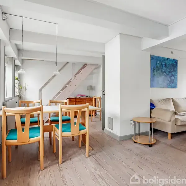 Spisebord med fire træstole med blå hynder står i et lyst rum med hvide vægge. Der er en trappe i baggrunden, en beige sofa til højre og et blåt maleri på væggen.