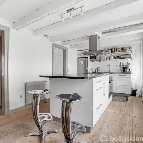 Køkken med en central køkkenø og to moderne barstole i et lyst rum med synlige loftsbjælker. Omkring er skabe, et komfur, og store vinduer, der giver naturligt lys.