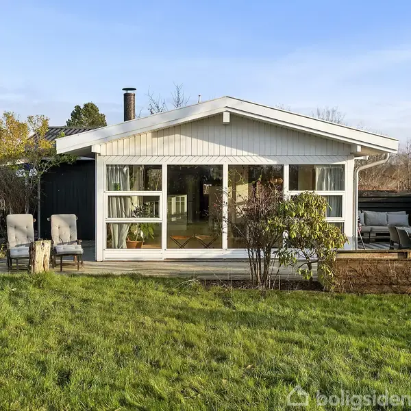 Et lille sommerhus med store vinduer vender ud mod en grøn græsplæne. To stole og en potteplante står på terrassen. Omkring er der buske og træer.