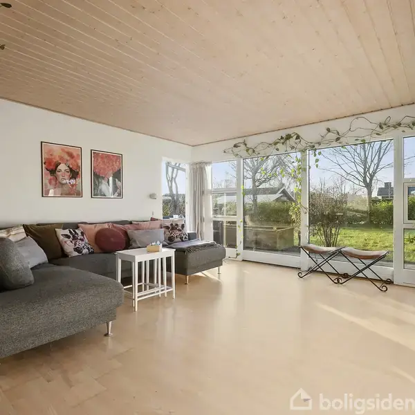 En grå sofagruppe står i en stue med lyse trægulve og træloft. Store vinduer fylder rummet med naturligt lys og giver udsigt til en have. Planter hænger ned fra loftet.