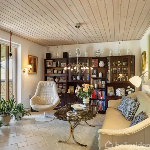 Et hyggeligt stueområde med en hvid lænestol og en beige sofa omkring et marmorbord med blomster. Bogreoler fyldt med dekorationer udgør baggrunden, og flere planter står ved vinduet.