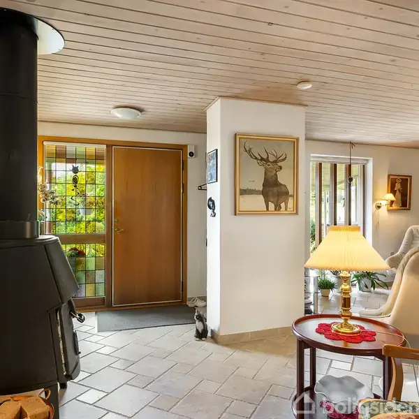 Brændeovn står ved siden af en trædør i en stue med lyst trægulv og vægge. Maleri af en hjort hænger på væggen, omgivet af komfortable stole og en lampe.