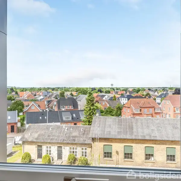Et vindue rammer en udsigt over en rolig bydel med flere hustage og træer, der strækker sig mod en blå himmel.