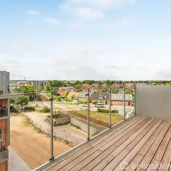 En træterrasse med glasrækværk strækker sig ud mod en udsigt over en by med huse og grønne områder under en let overskyet himmel.