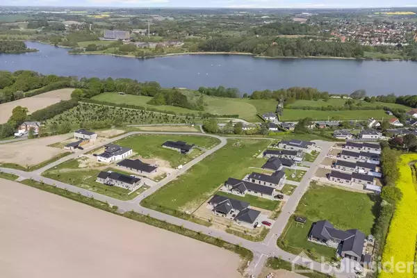 Huse under opførelse på en grøn byggeplads ved en sø i et landligt område med marker og skov i baggrunden.