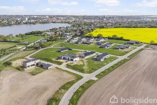 Boliger ligger spredt ud på grønne områder mellem asfaltveje. Foran ses en mark, mens en stor gul mark og en sø kan anes i baggrunden, nær en by.