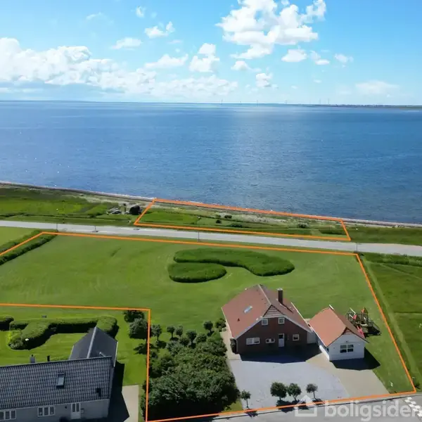 Et hus ligger ved en stor græsplæne med udsigt over havet. Rundt om er der markeret jordstykker, en vej skiller ejendommen fra kysten. Flere bygninger ses i baggrunden.