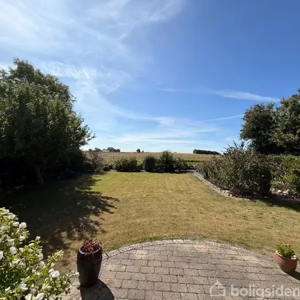 Græsplæne strækker sig fra en stenterrasse, omkranset af tætte buske og træer. Sollys oplyser området, mens en blå, næsten skyfri himmel ses i baggrunden.