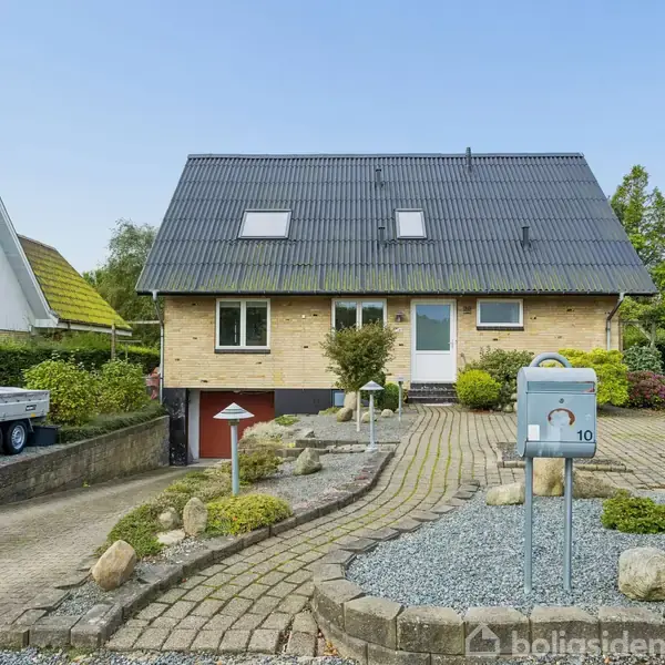Et murstenshus med skråt tag står placeret i en velplejet have med en flisebelagt indkørsel. Et postkassestativ markerer adressen med nummeret "10."