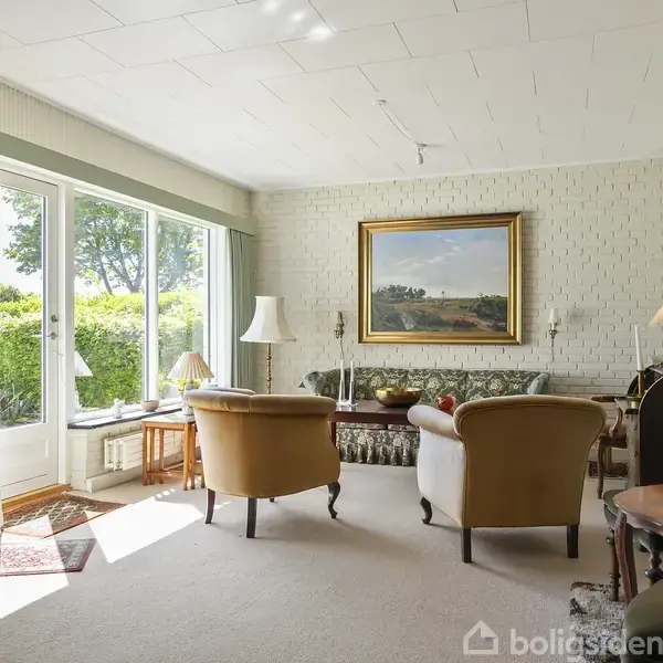 En stue med tre lænestole og en sofa omkring et sofabord. Væggene er dekoreret med malerier, og store vinduer giver udsigt til en have. Sollys strømmer ind, hvilket skaber en lys atmosfære.