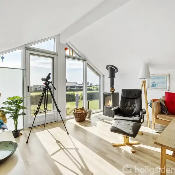 Stue med moderne indretning. En kikkert står på et stativ foran store vinduer med udsigt til naturen. En lænestol og sofa ved siden af pejsen, omgivet af planter.