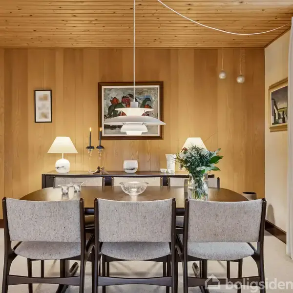 Et spisebord med fire stole står i et rum med trævægge og loft. En lampe hænger over bordet, og der er billeder og planter på en kommode i baggrunden.