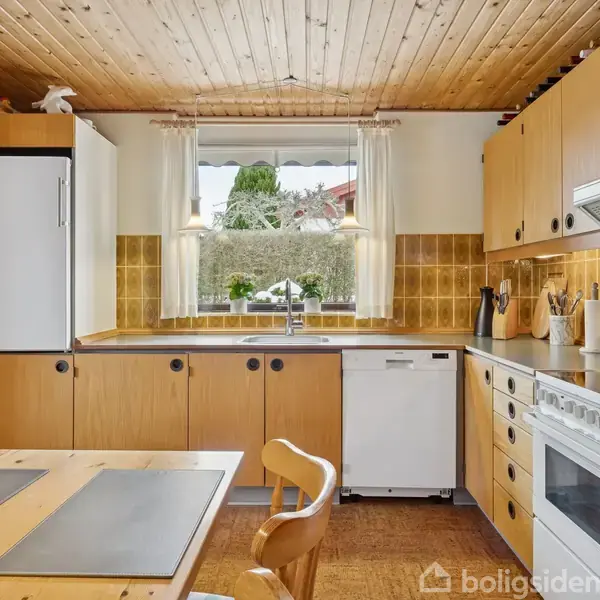 Et lyst køkken med træloft indeholder træmøbler og skabe, en hvid ovn og opvaskemaskine. Et stort vindue over vasken giver udsigt til en have.