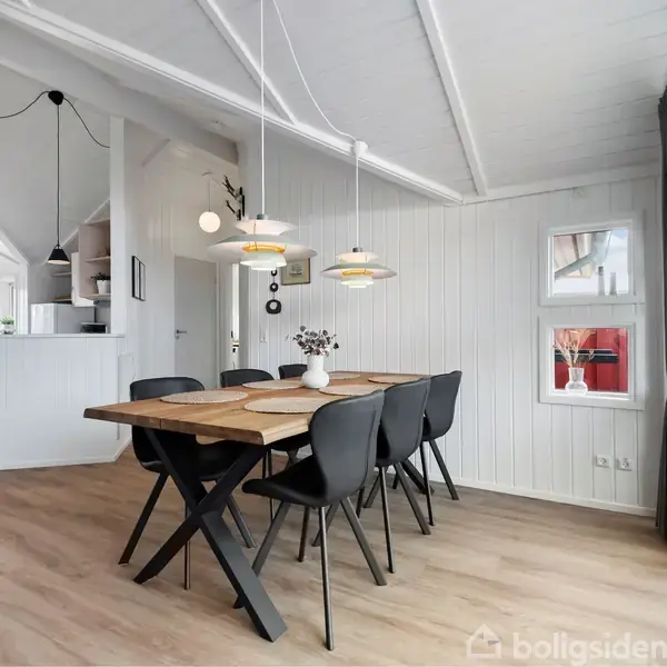 Et træbord med sorte stole står i en lys spisestue. Over bordet hænger moderne lamper. Rummet har hvidmalede vægge, store vinduer og træloft. Der er en åben adgang til køkkenet.