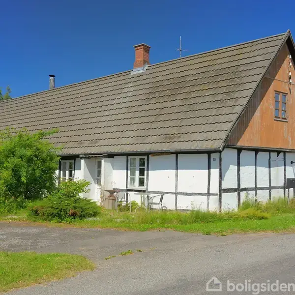 Et gammelt bindingsværkshus står stille i sollys, omgivet af grønne buske og græs. Huset har et gråt tag og en hvid facade med sorte bjælker.