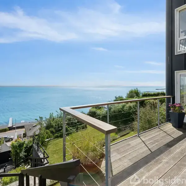 Balkon med træterrasse, der vender mod havet; omgivet af glasrækværk. I baggrunden ses frodige buske og en havudsigt med klart, blåt vand under en solrig himmel.