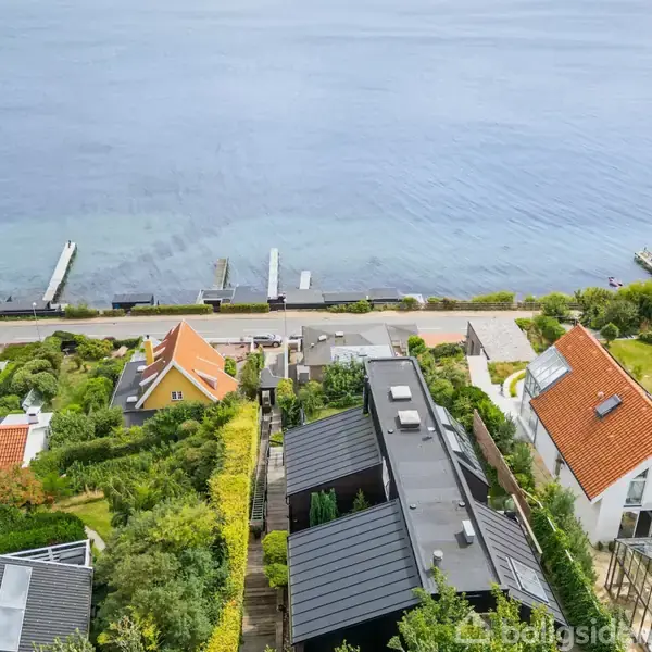 Huse med røde tage og haver ligger langs en vej ved vandet. Flere bådebroer strækker sig ud i vandet.