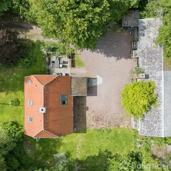 Et hus med rødt tag står ved siden af en gårdsplads omgivet af grønne træer og buske. En sti og en terrasseområde ses i nærheden.