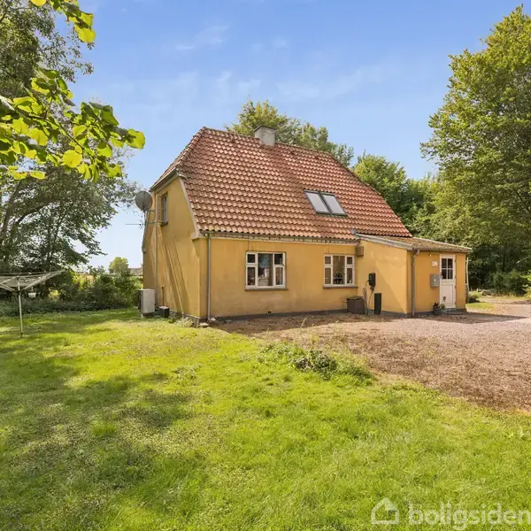 Gult hus står stille i en frodig have omgivet af træer og en grusbelagt indkørsel, med blå himmel i baggrunden.