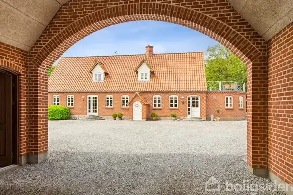 Rødt murstenshus med orange tegltag står stille i en gårdsplads omgivet af grus og grønne buske, set gennem en bueformet åbning.