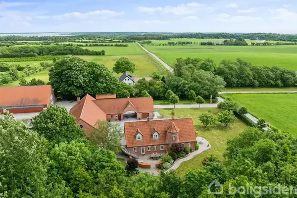 Et landsted med rødt tag ligger omgivet af grønne marker og træer. En grusvej fører til ejendommen, der har flere bygninger og en frodig have.