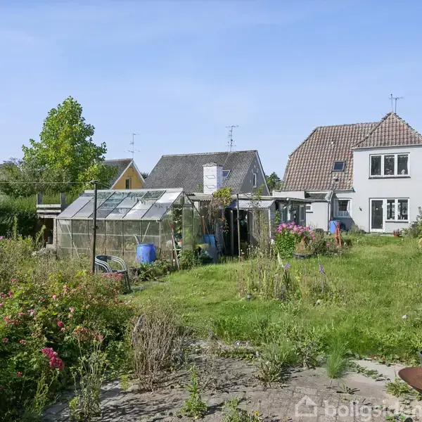 Et lille hus med hvide vægge og brunt tag står i en have med græs, buske og et drivhus. Omgivet af andre huse i en villakvarter.