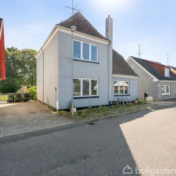 Grå murstenshus med to etager står på en rolig villavej. Omgivet af andre farverige huse under en klar blå himmel.