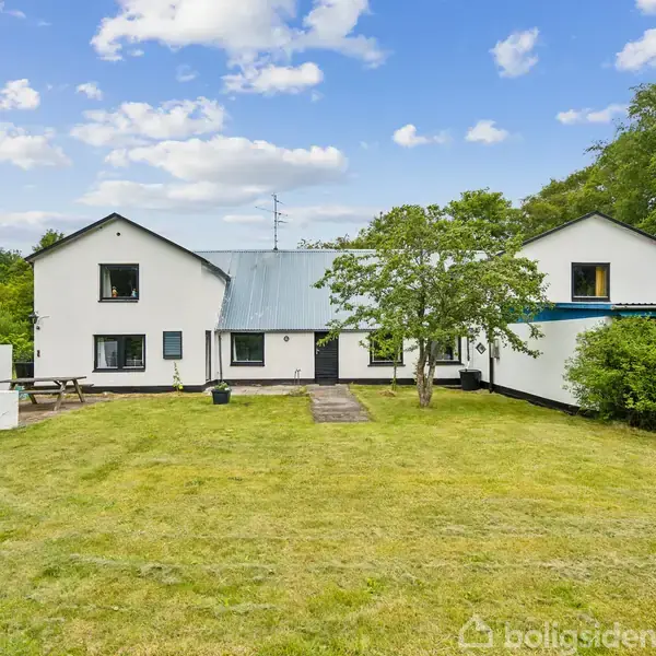 Et hvidt to-etagers hus med mørke vinduesrammer står stille i en grøn have omkranset af træer. En græsplæne strækker sig foran med et par buske og et træ.