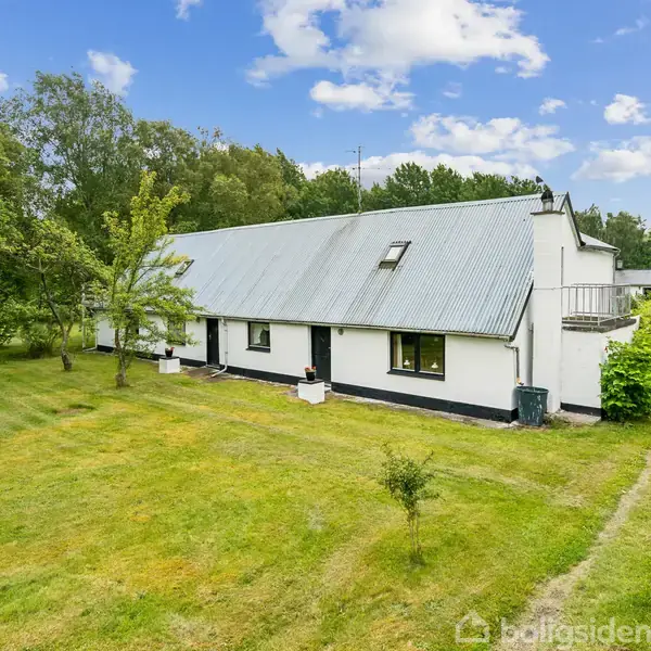 Hus med hvid facade og gråt tag står stille omgivet af en stor græsplæne og træer under en blå himmel med skyer.