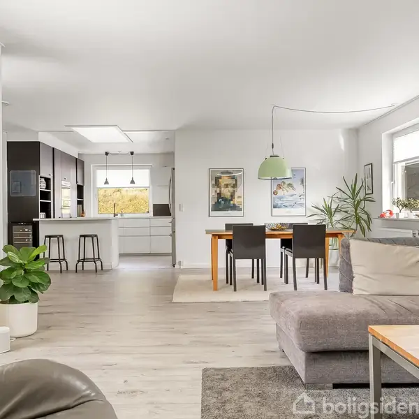 En lys, åben stue med hjørnesofa og træ sofabord, der vender mod et åbent køkken. Spisebordet har seks stole, omgivet af vinduer og grønne planter.
