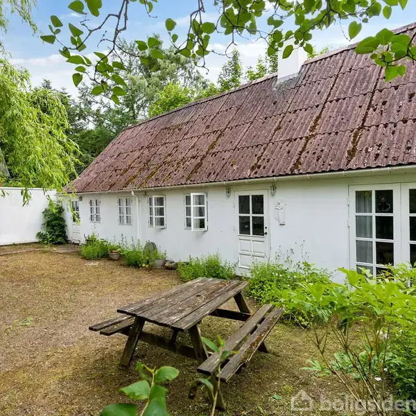 Et hvidt hus med rødt tag står i en have med en træbænk og bord i forgrunden. Omkring er der grønne buske og træer, der skaber en rolig atmosfære.