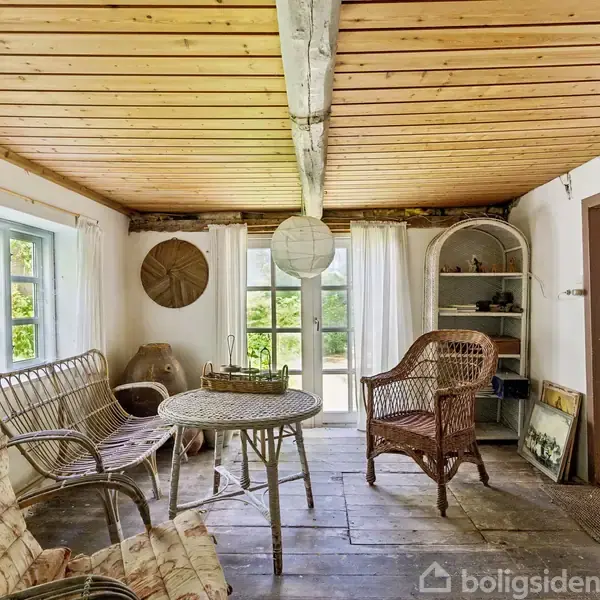 En rustik stue med trægulv og træmøbler, herunder en sofa og stole. Et rundt bord er centralt placeret. Rummet er lyst med store vinduer og en åben dør.