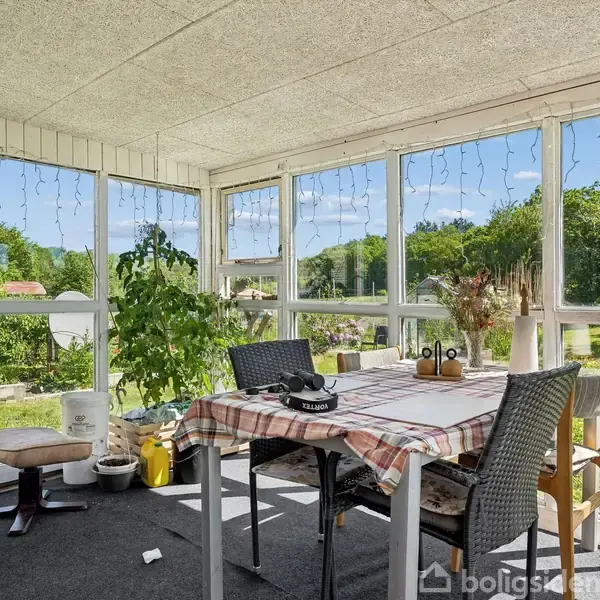 Et bord med ternet dug står i en lys udestue omringet af stole. Planter og pyntegenstande er arrangeret omkring. Udenfor vinduerne ses en grøn have og blå himmel.