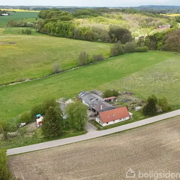 Længere vej krydser en række bygninger og marker omgivet af skovklædte områder i et frodigt, landligt landskab med rullende bakker.
