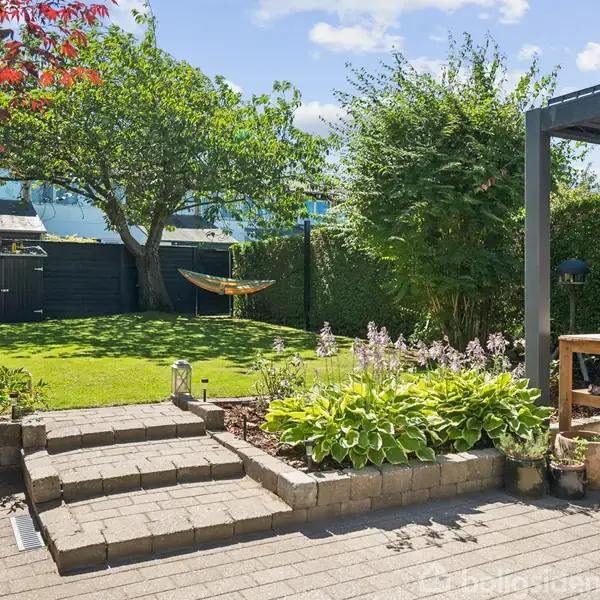 Grøn have med en stor græsplæne og træskur i baggrunden. Forgrund har en terrasse med fliser, planter og en grill under et halvtag. Solskin oplyser området.