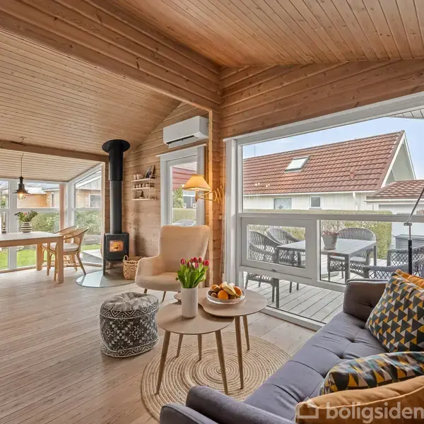 Stue med en sofa og bord med blomster og frugter. Brændeovn ved siden af stol. Store vinduer med udsigt til terrasse og hus. Lys indretning med trægulve og trævægge.