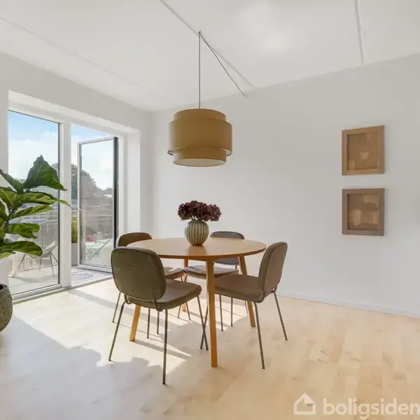 Rundt spisebord med fire stole og en vase med blomster på bordet. Store vinduer åbner til balkon; til venstre står en stor plante, og til højre ses en køkkenbord med to barstole.