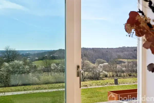 En glasdør står åben, hvilket giver udsigt til en grøn have og skovklædte bakker i baggrunden under en klar blå himmel. En vase med tørrede blomster er i forgrunden.