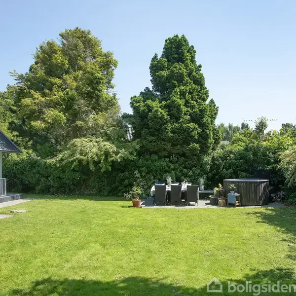En velplejet græsplæne med havemøbler står på en terrasse. Til venstre ses en del af et hus. Omgivet af frodig, tæt beplantning.