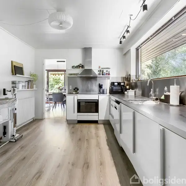 Et moderne køkken med hvidt interiør har komfur og emhætte. Store vinduer med udsigt til frodigt grønt. Rummets lyse trægulv fører til en spisestue.