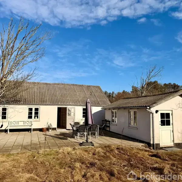 Et hvidt hus med gråt tag står stille. Terrassen har et bord med stole og en parasol. Træer uden blade omkranser huset under en blå himmel.